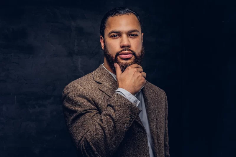 Confident african-american businessman in a brown classic jacket, thinking about business while holding hand on chin. isolated on a dark background. Free Photo