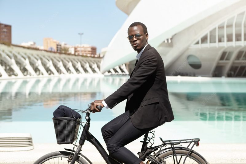 Confident african american businessman in formal wear commuting to work on black bicycle. corporate worker hurrying to office on bike. eco-friendly transport and healthy active lifestyle concept Free Photo