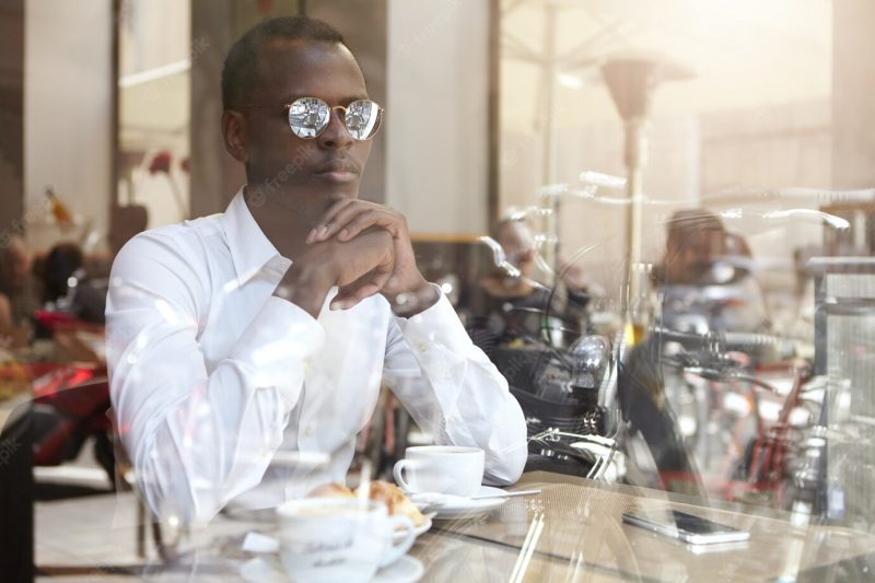 Confident thoughtful young African American businessman in stylish eyewear clasping hands while sitting at coffee shop with mug on table, came to think of business issues over cup of cappuccino Free Photo