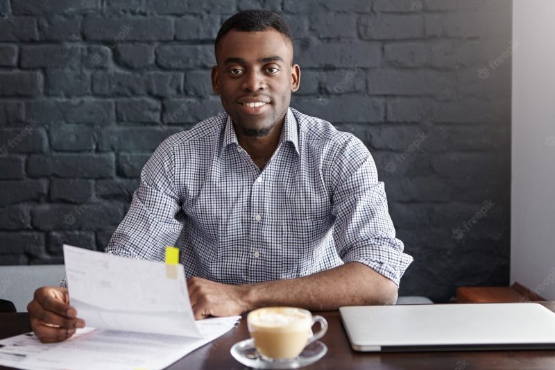 Confident young African businessman wearing formal shirt sitting at table with laptop, mug and papers Free Photo