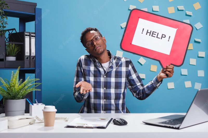 Confused and clueless company office worker holding help cardboard bubble sign while shrugging in workspace. bewildered uncertain agency employee raising shoulders ambiguous. Free Photo