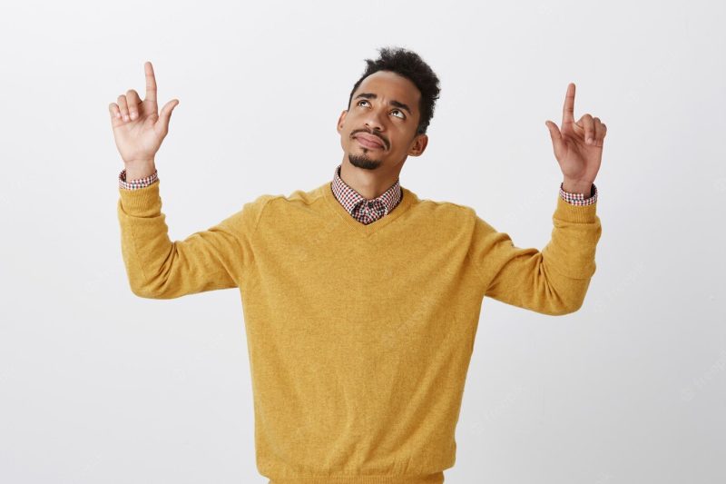 Confused guy, having trouble to recognize something upstairs. questioned beautiful dark-skinned male model with afro hairstyle, raising hands, pointing and looking up with doubts and suspicion Free Photo