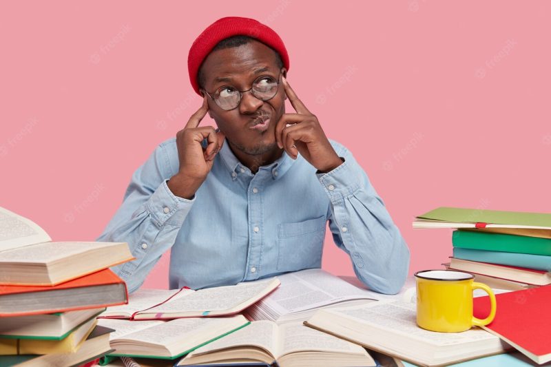 Contemplative dark skinned college student keeps both fingers on temples, purses lips and looks with hesitaton aside, tries analyze information in his mind, being workaholic Free Photo