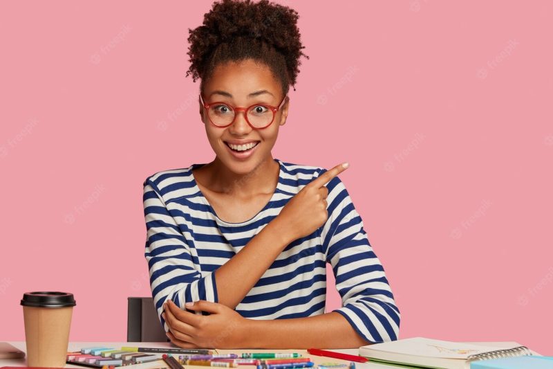 Content black craftswoman in striped clothes, shows free space against pink wall, works on new sketch in notebook with crayons, drinks take out coffee, works from home, has creative skills Free Photo