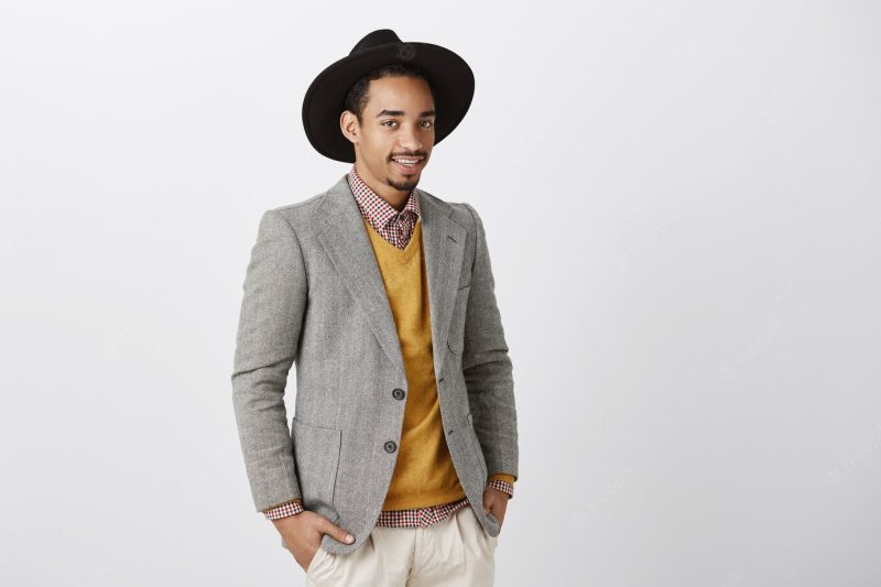 Creative designer discussing fashion show. good-looking dark-skinned male model in stylish jacket and hat, standing half-turned, talking casually with coworkers about work and lifestyle over gray wall Free Photo