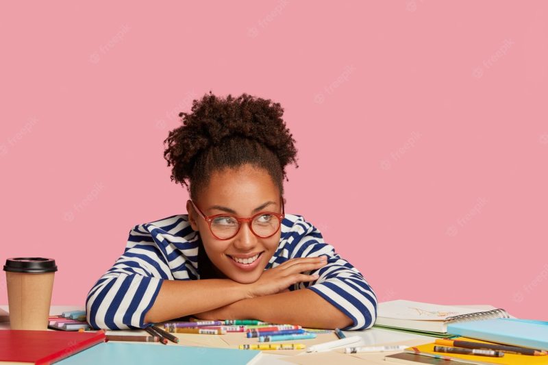 Creativity and inspiration concept. pleased smiling afro american graphic designer leans at table, has coffee break after drawing picture in notebook, isolated over pink wall in art Free Photo