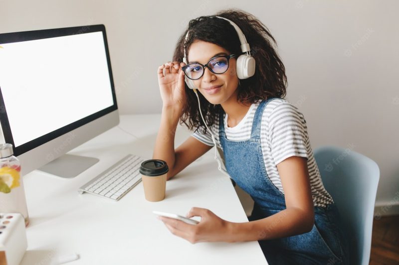 Cute girl with tired smile posing at workplace near computer with white screen Free Photo