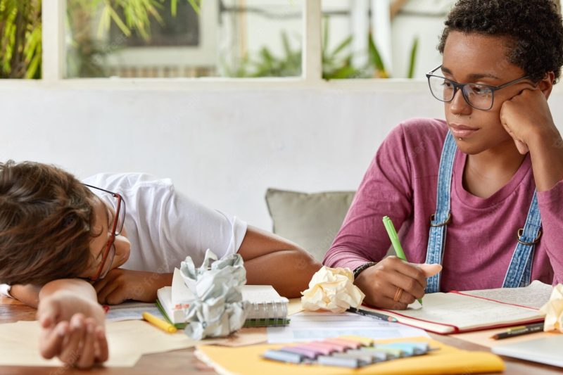 Dark skinned female student wears transparent glasses, looks seriously at tired classmate, work together at course paper, pose at desk with papers and notepad, collaborate to learn material. Free Photo