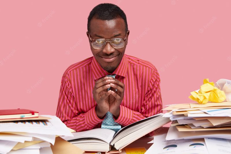 Dark skinned pleased African American college student keeps hand in intriguing gesture, has nice idea to express, reads thick book Free Photo