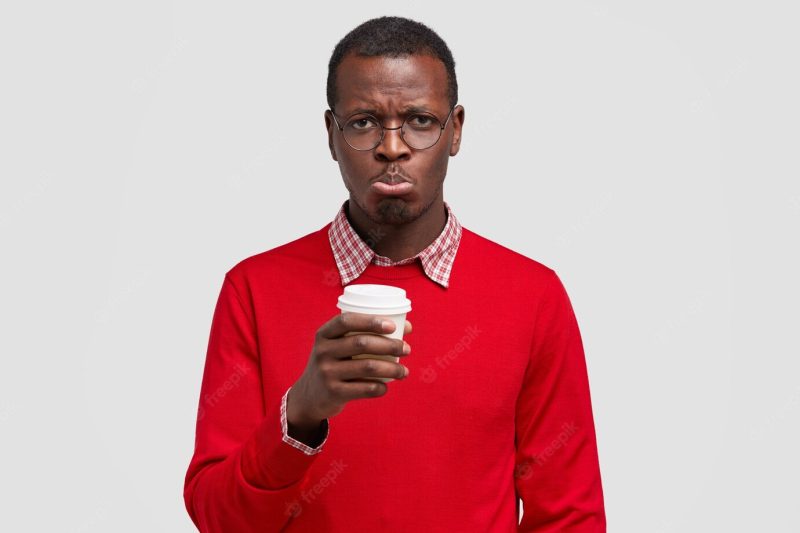 Dejected dissatisfied guy has dark skin, miserable facial expression, purses lips in discontent, drinks take out coffee Free Photo