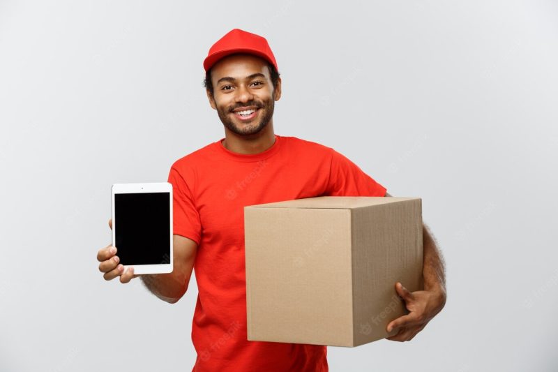 Delivery concept – portrait of handsome african american delivery man or courier with box showing tablet on you to check the order. isolated on grey studio background. copy space. Free Photo