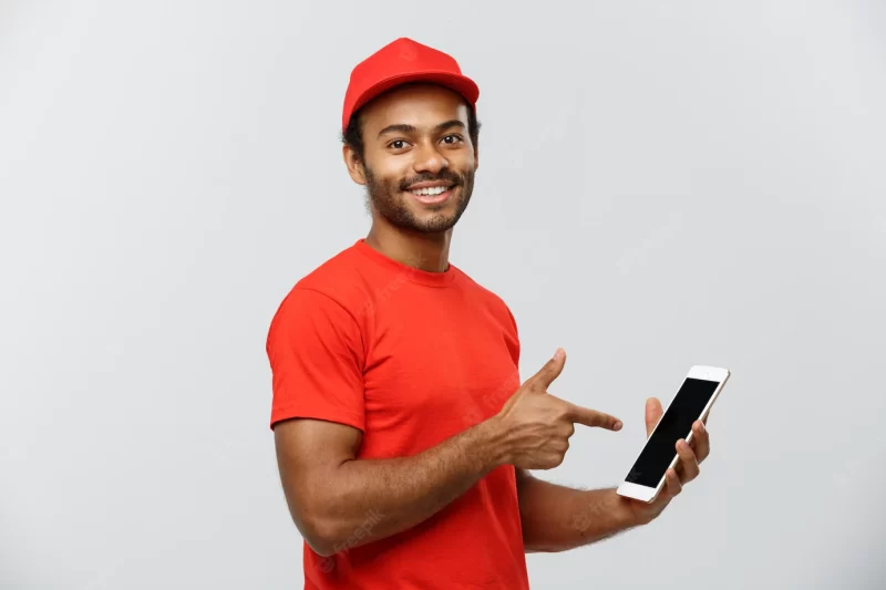 Delivery concept – portrait of handsome african american delivery man or courier with box showing tablet on you to check the order. isolated on grey studio background. copy space. Free Photo