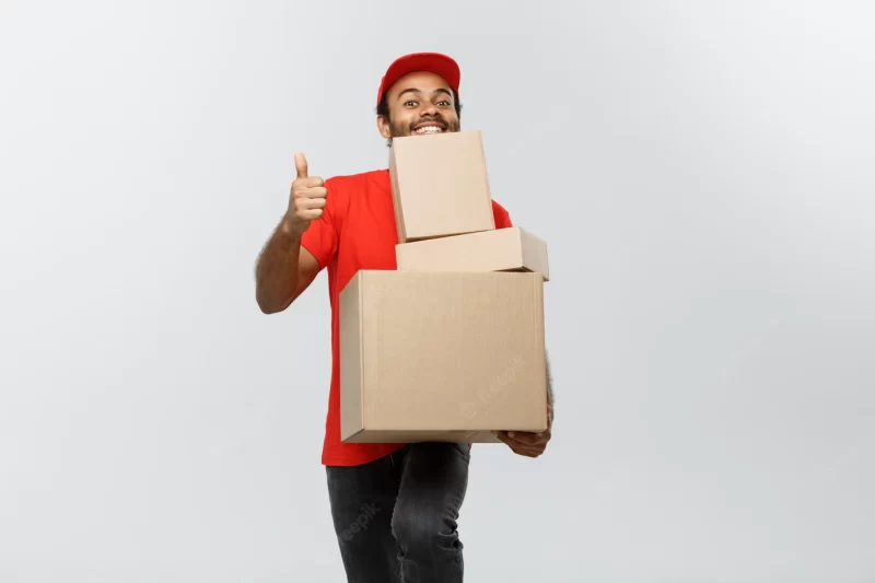 Delivery concept – portrait of happy african american delivery man in red cloth holding a box package. isolated on grey studio background. copy space. Free Photo