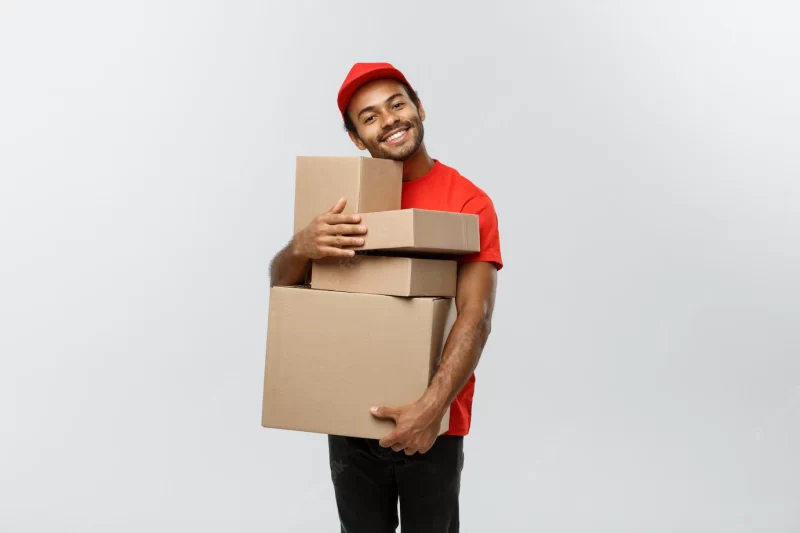 Delivery concept – portrait of happy african american delivery man in red cloth holding a box package. isolated on grey studio background. copy space. Free Photo