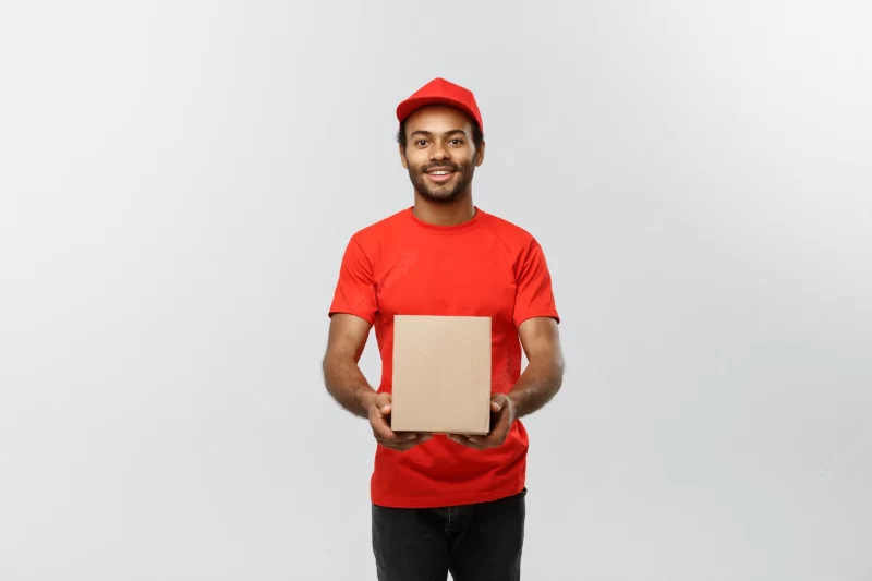 Delivery concept – portrait of happy african american delivery man in red cloth holding a box package. isolated on grey studio background. copy space. Free Photo