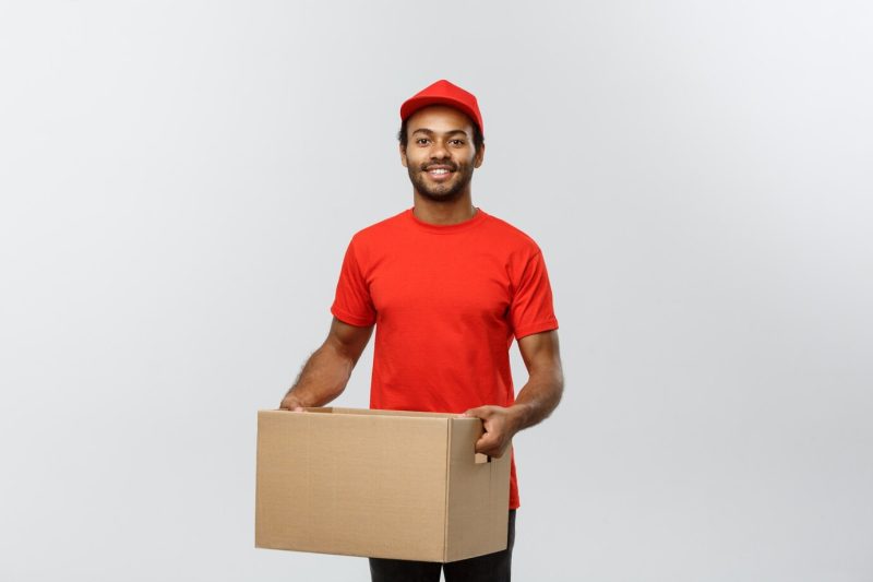 Delivery concept – portrait of happy african american delivery man in red cloth holding a box package. isolated on grey studio background. copy space. Free Photo