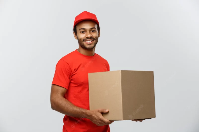 Delivery concept – portrait of happy african american delivery man in red cloth holding a box package. isolated on grey studio background. copy space. Free Photo