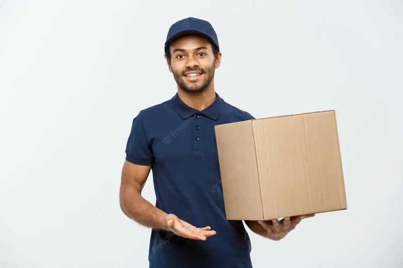 Delivery concept – portrait of happy african american delivery man in red cloth holding a box package. isolated on grey studio background. copy space. Free Photo