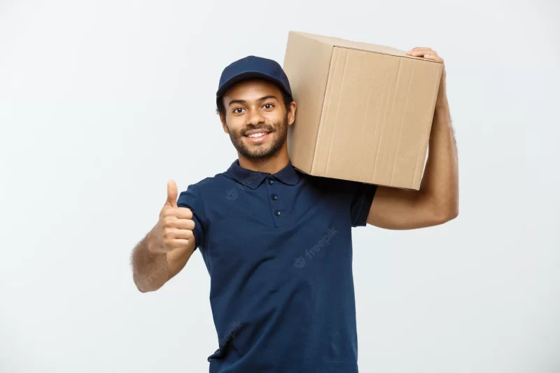 Delivery concept – portrait of happy african american delivery man in red cloth holding a box package. isolated on grey studio background. copy space. Free Photo