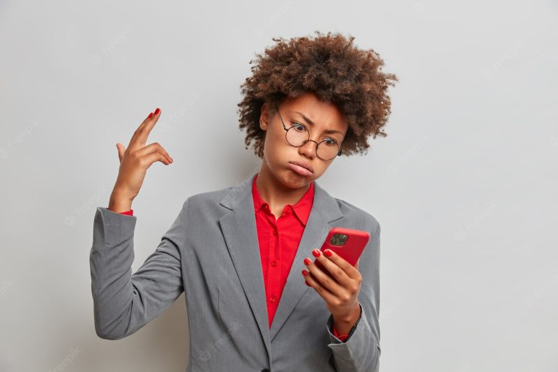 Desperate exhausted curly female corporate worker makes finger gun pistol, being sick and tired of receiving messages from stranger, holds modern cellular, wears business outfit Free Photo