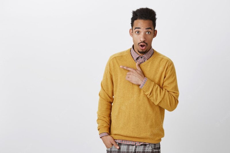 Discussing with friend shocking gossip about classmate. attractive African-American male student with Afro hairstyle in yellow pullover, dropping jaw from surprise, pointing at upper l