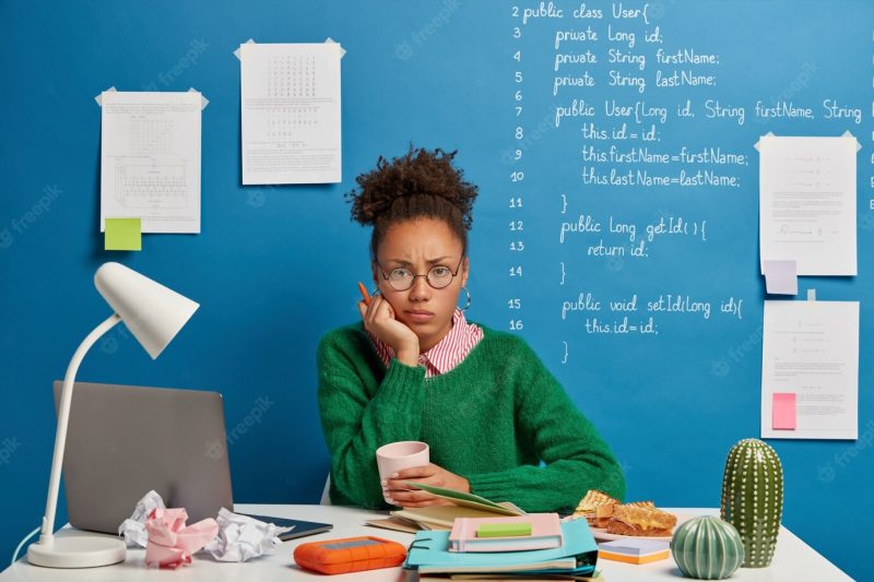 Displeased afro woman holds pen, feels tired of exam preparation, drinks coffee and looks sadly at camera, sits at home desk with necessary equipment and notepad Free Photo