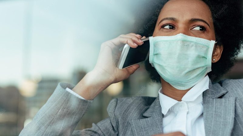 Distraught black businesswoman communicating over smart phone while wearing face mask during virus epidemic the view is through the glass Free Photo