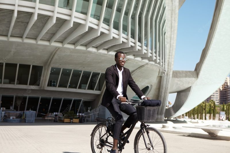 Ecologically conscious young black european businessman wearing black suit and sunglasses cycling to cafe on retro bicycle during lunch break on sunny day Free Photo