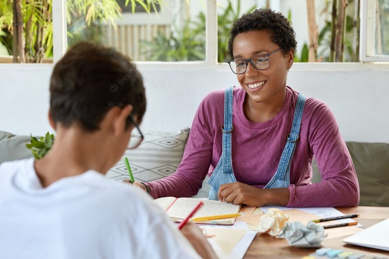 Education, relationship and collaboration concept. satisfied black young woman with curly hair, wears spectacles for vision correction, looks positively at pupil who tries understand material Free Photo