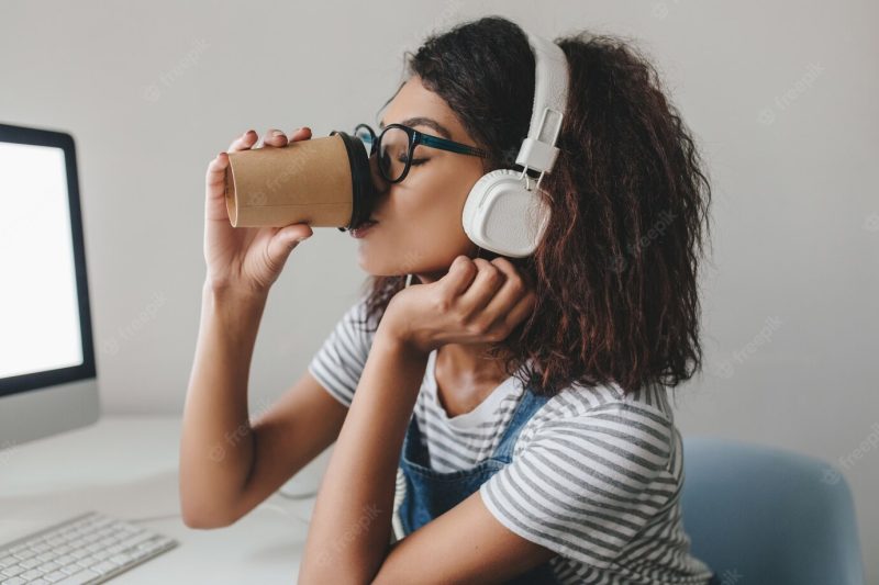 Elegant black girl with curly hairstyle drinking coffee at workplace posing near gray wall Free Photo