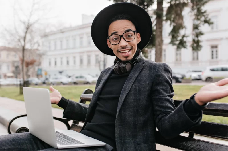 Emotional african student in glasses sitting on bench with laptop. outdoor photo of mulatto male freelancer in black attire working with computer. Free Photo