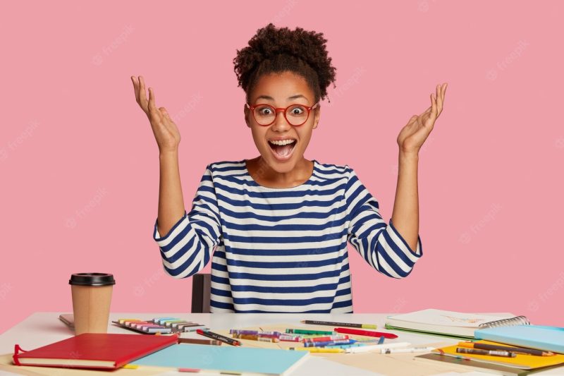 Emotional illustrator raises hands in eureka, exclaims with happiness, has nice idea for masterpiece, works at table with notepad, crayons, coffee, wears striped sweater, isolated over pink wall Free Photo