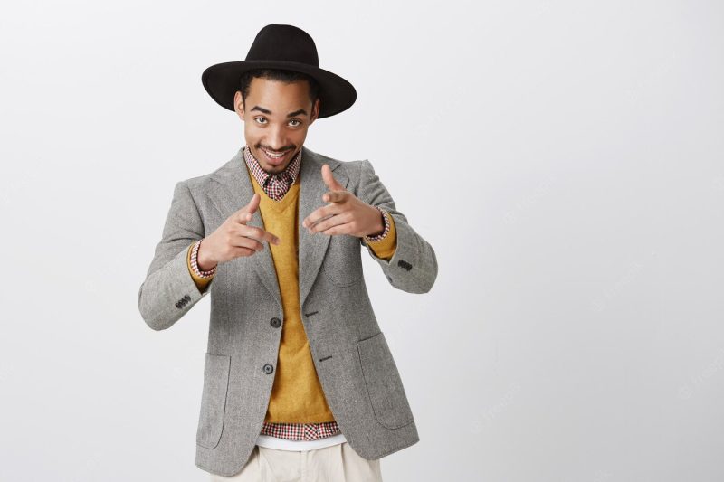 Emotive party guy inviting us to club. portrait of positive good-looking businessman in stylish hat and jacket, flirting or being macho, pointing with gun gesture, standing over gray wall Free Photo