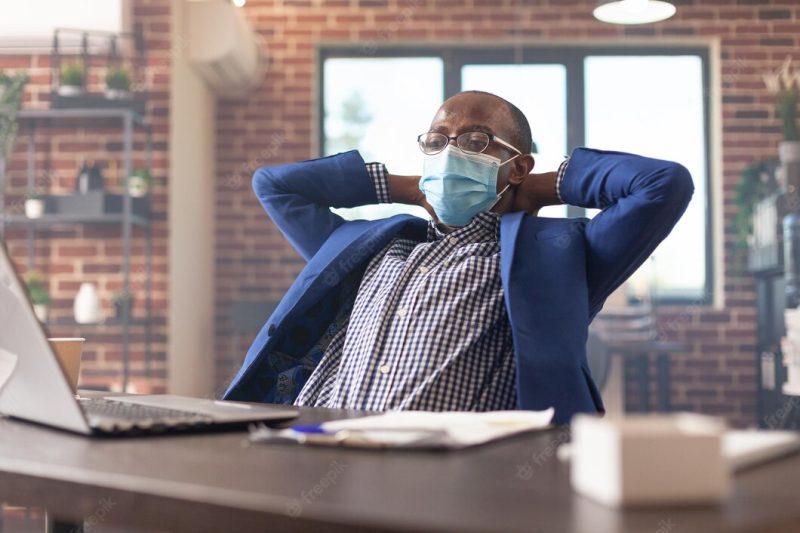 Employee relaxing in office after finishing business project, wearing face mask. calm entrepreneur taking break to relax after work and getting job done. man sitting with hands over head. Free Photo