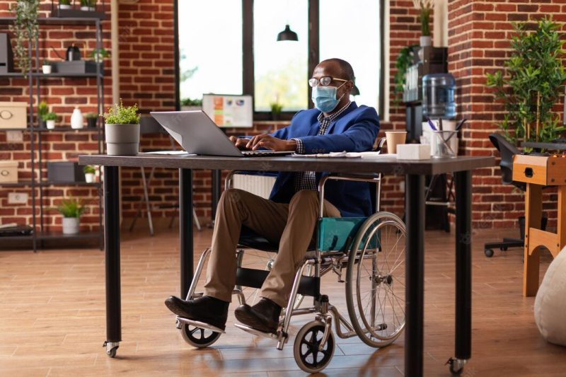 Employee with disability wearing face mask and working on business project. entrepreneur sitting in wheelchair, doing financial and marketing work during coronavirus pandemic. man at job Free Photo