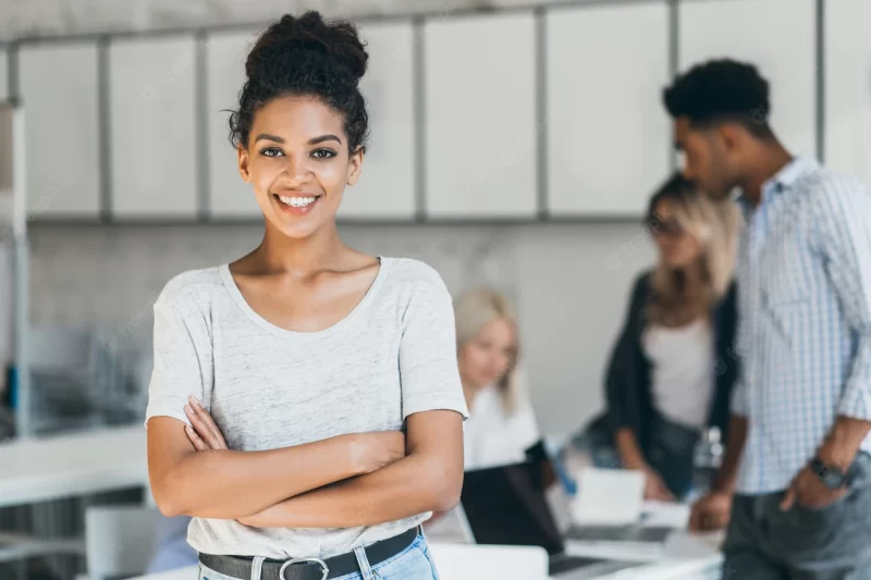 Enthusiastic female african student enjoying company with friends in lecture hall. indoor portrait of smiling black office worker posing with arms crossed in front of foreign colleagues. Free Photo