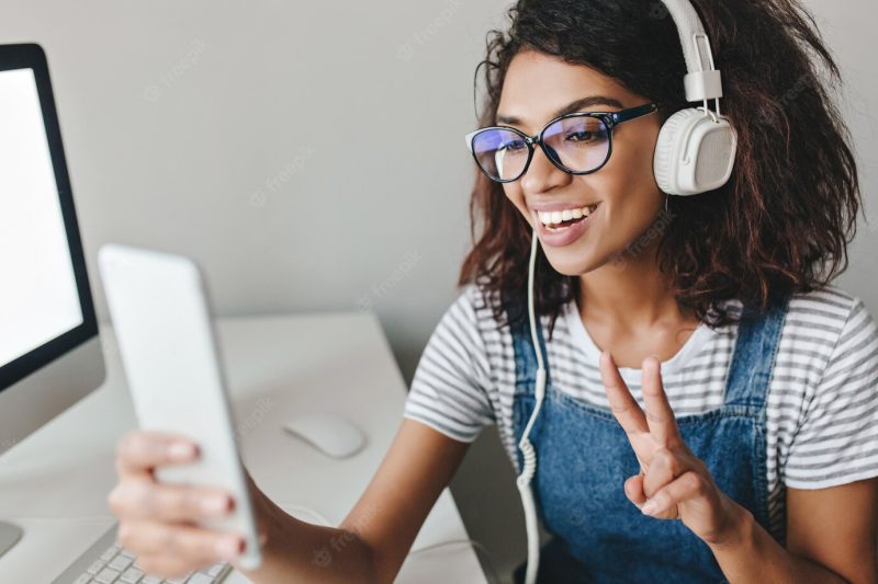 Enthusiastic girl wearing glasses making selfie with peace sign spending time in office Free Photo