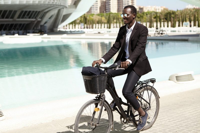 Environmentally friendly afro american banker in formal wear and shades looking happy and relaxed, cycling to work on bike in urban setting, smiling cheerfully. business people, lifestyle and transport Free Photo