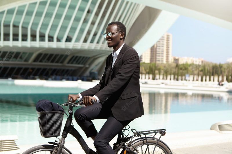 Environmentally friendly black manager in formal suit and sunglasses looking forward while riding bicycle to work in urban surroundings, smiling happily. business, lifestyle, transport and people Free Photo