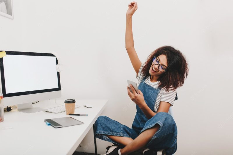 Excited black girl sitting with legs crossed and happy laughing, reading phone message Free Photo