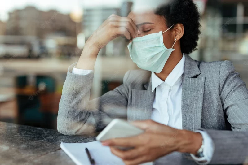 Exhausted african american businesswoman wearing protective face mask and holding her head in pain the view is through the glass Free Photo