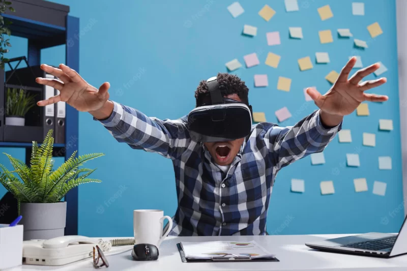 Expressive amazed african american company office employee enjoying virtual reality gaming technology. adult businessman wearing electronic entertainment device while sitting at desk in workspace. Free Photo