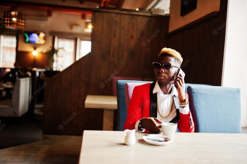 Fashion african american man model at red suit with highlights hair and sunglasses sitting at cafe with coffee and speaking on mobile phone Free Photo