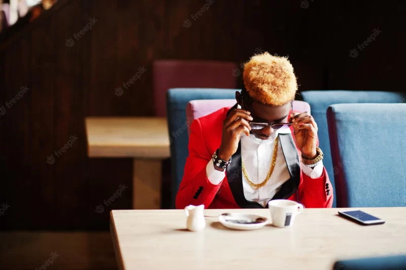 Fashion african american man model at red suit with highlights hair and sunglasses sitting at cafe with coffee and speaking on mobile phone Free Photo