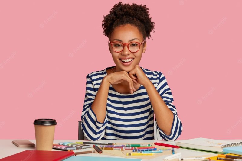 Fashion designer with toothy smile, feels sarisfied, keeps hands under chin, wears striped clothes, isolated over pink wall. positive painter has inspiration for drawing. creative work concept Free Photo