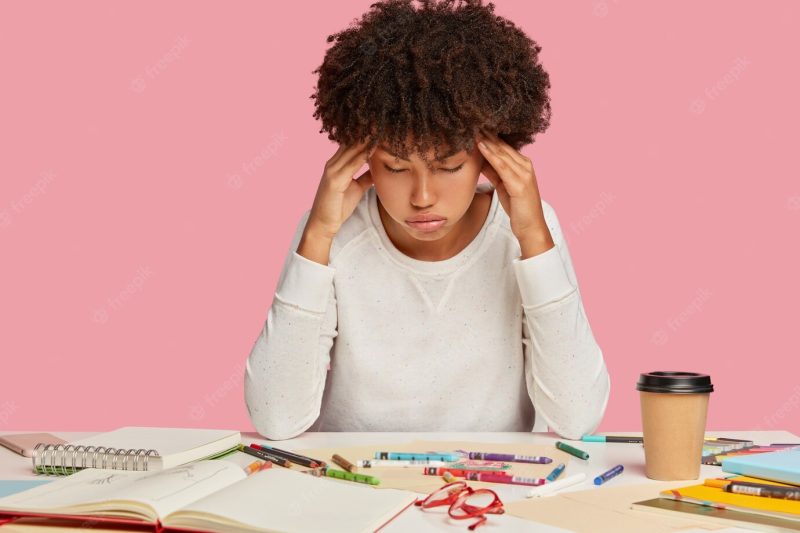 Fatigue african american woman keeps hands on temples, suffers from migraine, sighs with tiredness, works for long time, poses at desk with spiral notepad, takeaway coffee, isolated on pink wall Free Photo