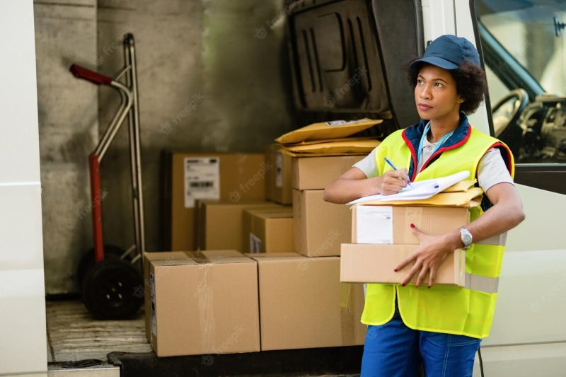 Female African American courier getting ready for the delivery and checking package list by delivery van Free Photo