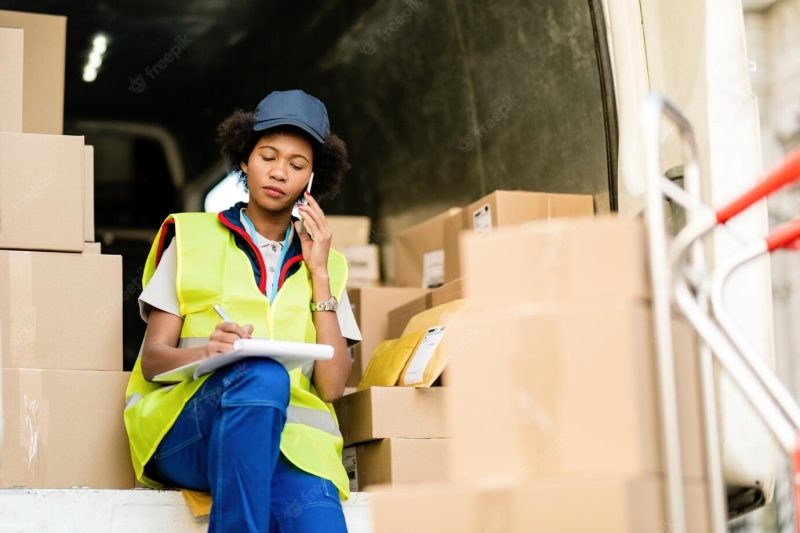 Female black courier sitting in delivery van and communicating on mobile phone while writing notes in clipboard Free Photo