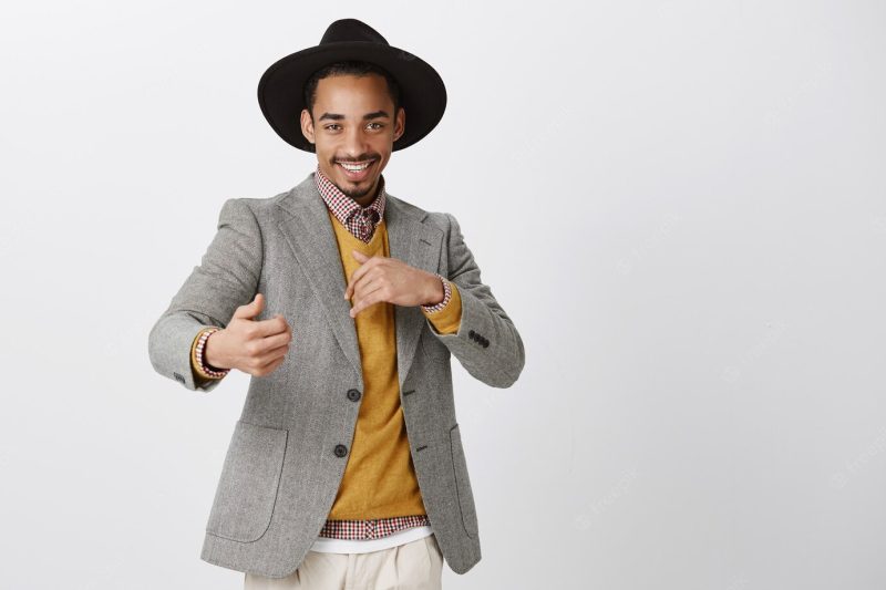 Flirty romantic male wanting have fun. satisfied confident African-American man in stylish black hat and jacket, making sensual and passionate gestures, smiling curiously over gray wall Free Photo