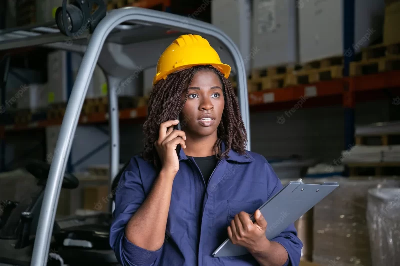 Focused black warehouse worker in yellow hardhat standing near forklift and talking on cell. shelves with goods in background. medium shot. labor or communication concept Free Photo